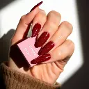 High contrast, low-key lighting, natural earth tones. Close-up of a hand with short almond-shaped nails, deep ruby red with sparkles and velvet finish, holding a pink Glamnetic nail box. White or blush pink backgro.webp