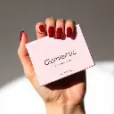 A close-up, high-resolution image of an adorable hand with short, almond-shaped, deep ruby red press-on nails holding a soft pink Glamnetic nail box. The background is clean white. Contrasting chiaroscuro lighting .webp
