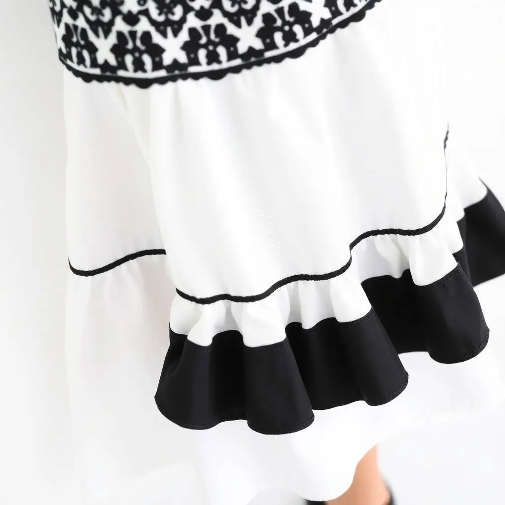 Close-up shot of a sleeveless white cotton poplin dress with black embroidered yoke, fabric belt, and ruffle tiers, on a white background.  Fit-and-flare silhouette, round neckline, styled with black ankle-strap he.webp