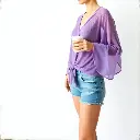 A cast bronze surface with a smooth gradient transition, depicting a woman in a lavender purple sheer mesh shrug with long bell sleeves and a tie-front, wearing distressed denim shorts and delicate rings, holding a.webp